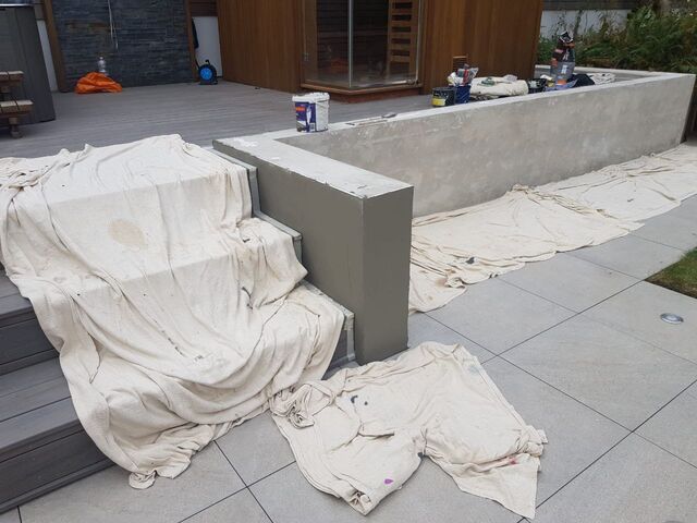 Dust sheets over grey paving and decking beside a rendered plinth, paint kettle and brushes on the wall