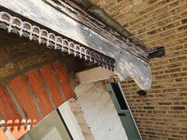 Stripped grey timber fascia with flaking paint and a pierced metal trim above a red brick arch