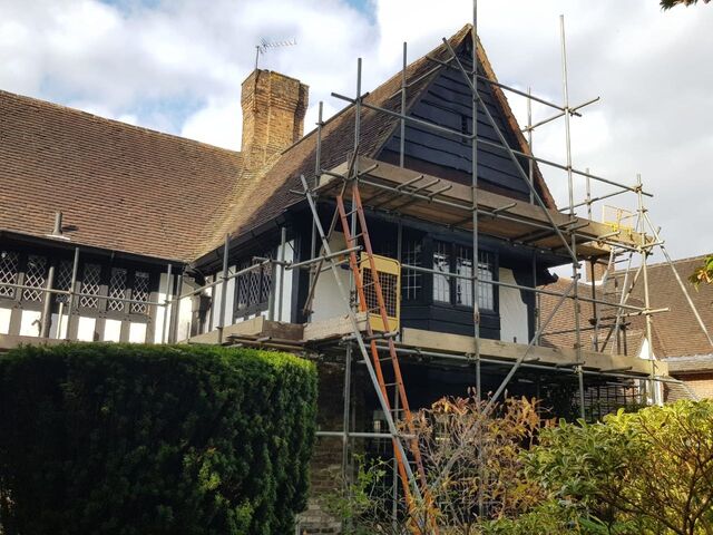 Black and white half-timbered house under scaffold, dark boarded gable above, ladder and hedge