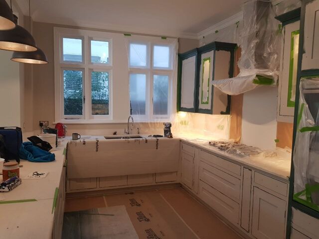Kitchen with dark green painted unit frames and white panels, worktops sheeted, sink below the window