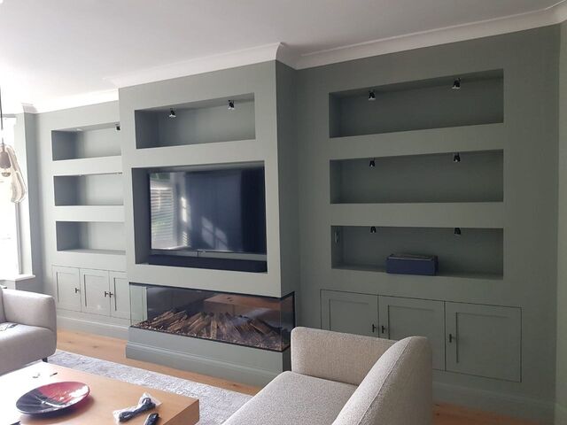 Living room media wall and alcove shelving painted pale grey, with a TV recess, log fire and cupboards