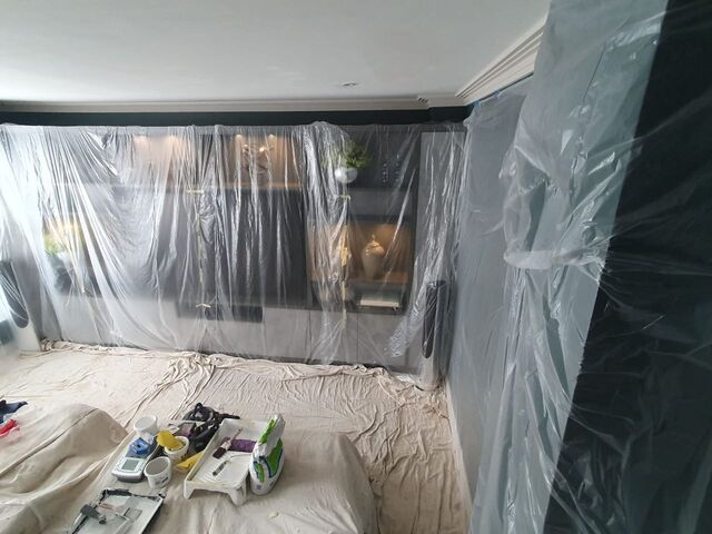 Plastic sheeting over a dark green painted wall with tools and paint kettles on sheets on the floor